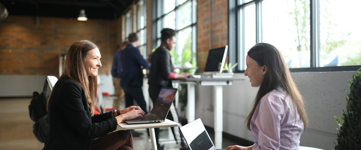 Two colleagues talking across their laptops in a bright, open brick-walled office.